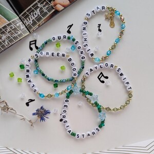 May include: Several beaded bracelets with white letter beads spelling out phrases like "Wishing Star" and "Taylor Swift." The bracelets feature green, blue, and gold accents, with musical note charms and scattered decorative beads.