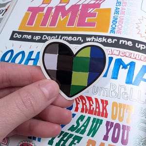 May include: A heart-shaped sticker with a color block design in shades of black, white, green, and blue. The sticker is held in a hand, with a background of colorful text including the word "TIME" and phrases like "FREAK OUT" and "SAW YOU".