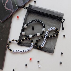 Several Taylor Swift friendship bracelets on a black organza gift bag on a white background. The bracelets have lyrics and titles from Taylor Swift's album Reputation including Delicate and Getaway Car.