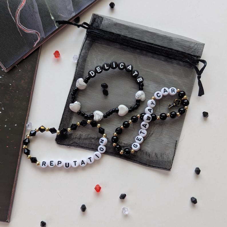 Several Taylor Swift friendship bracelets on a black organza gift bag on a white background. The bracelets have lyrics and titles from Taylor Swift's album Reputation including Delicate and Getaway Car.
