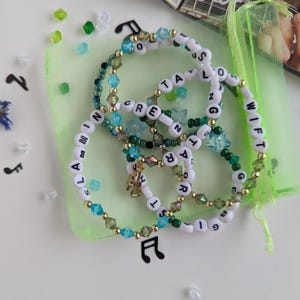 Several Taylor Swift friendship bracelets on a green organza gift bag on a white background. The bracelets have lyrics and titles from Taylor Swift's debut album including Slamming screen door, Taylor swift, Georgia stars, and more.