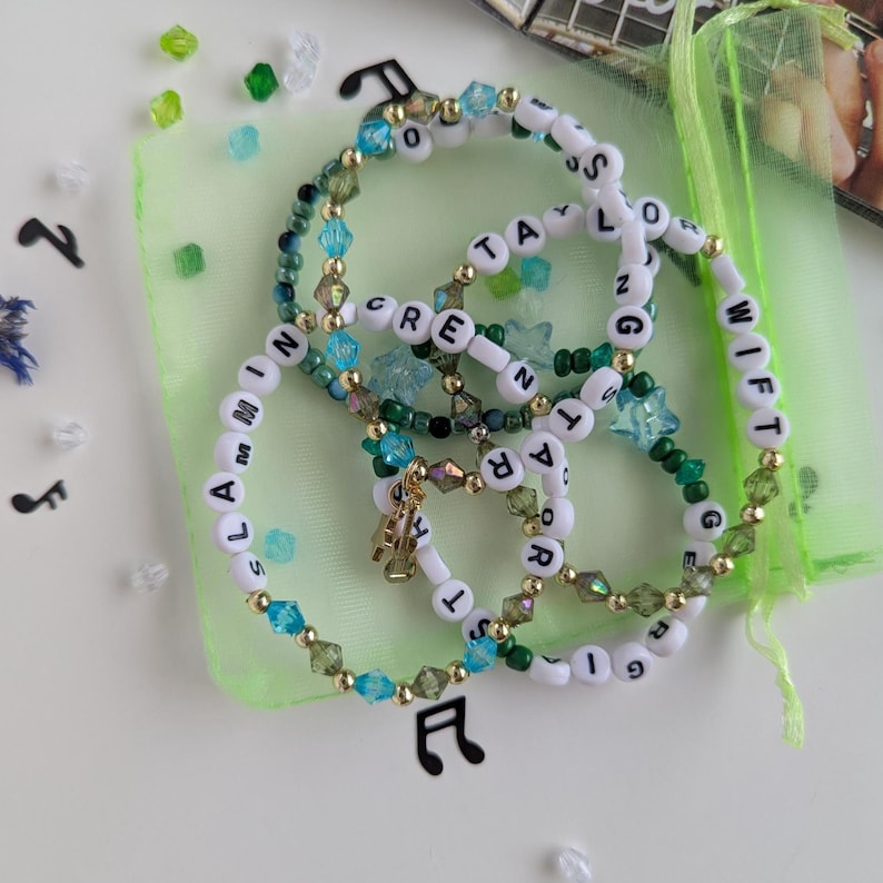Several Taylor Swift friendship bracelets on a green organza gift bag on a white background. The bracelets have lyrics and titles from Taylor Swift's debut album including Slamming screen door, Taylor swift, Georgia stars, and more.