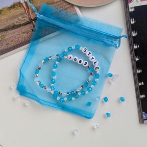 Several Taylor Swift friendship bracelets on a blue organza gift bag on a white background. The bracelets have lyrics and titles from Taylor Swift's album 1989 including 1989 and Style.