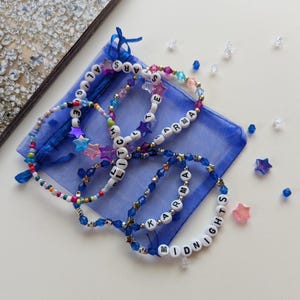 Several Midnights themed Taylor Swift friendship bracelets on a blue organza gift bag, featuring songs and lyrics from the Midnights album including Karma, Midnights, Glitch, All the stars aligned.