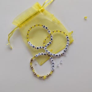 Several Taylor Swift friendship bracelets on a yellow organza gift bag on a white background. The bracelets have lyrics and titles from Taylor Swift's album Fearless including Love Story and You Belong with Me.