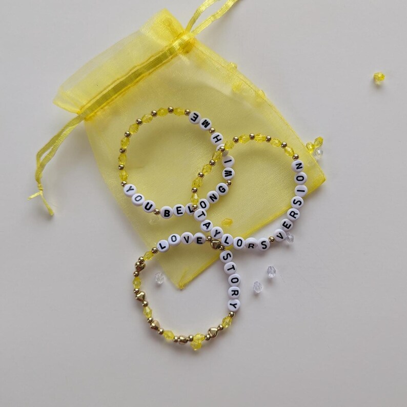 Several Taylor Swift friendship bracelets on a yellow organza gift bag on a white background. The bracelets have lyrics and titles from Taylor Swift's album Fearless including Love Story and You Belong with Me.