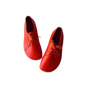 May include: A pair of vibrant red lace-up shoes featuring a smooth, rounded toe design and thin matching red laces. The shoes have a low-cut silhouette with a clean, minimalist construction and a solid, uniform color throughout the entire exterior.