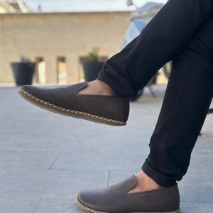 May include: A pair of brown leather slip-on shoes with a stitched edge detail. The shoes are worn by a person sitting with their legs crossed.