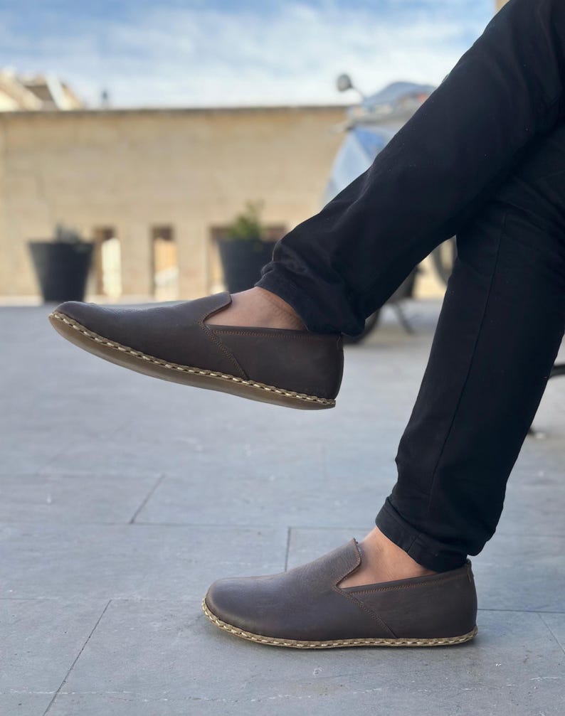 May include: A pair of brown leather slip-on shoes with a stitched edge detail. The shoes are worn by a person sitting with their legs crossed.
