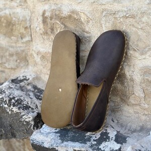 May include: A pair of brown leather slip-on shoes with a brown leather sole. The shoes are on a rock surface.