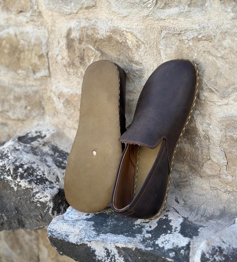 May include: A pair of brown leather slip-on shoes with a brown leather sole. The shoes are on a rock surface.