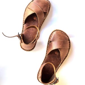 Op de afbeelding: Twee bruine leren sandalen met een kruisontwerp over de voet en een enkelband met een gesp. De sandalen hebben witte stiksels rond de randen en staan tegen een witte achtergrond.