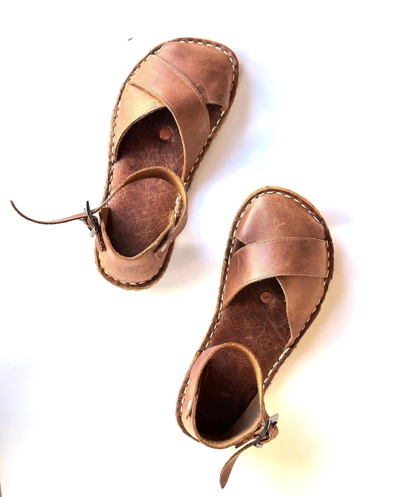Op de afbeelding: Twee bruine leren sandalen met een kruisontwerp over de voet en een enkelband met een gesp. De sandalen hebben witte stiksels rond de randen en staan tegen een witte achtergrond.