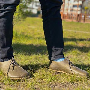 May include: A pair of olive green leather lace-up shoes with a rounded toe and a thick sole. The shoes are worn by a person wearing dark blue jeans.