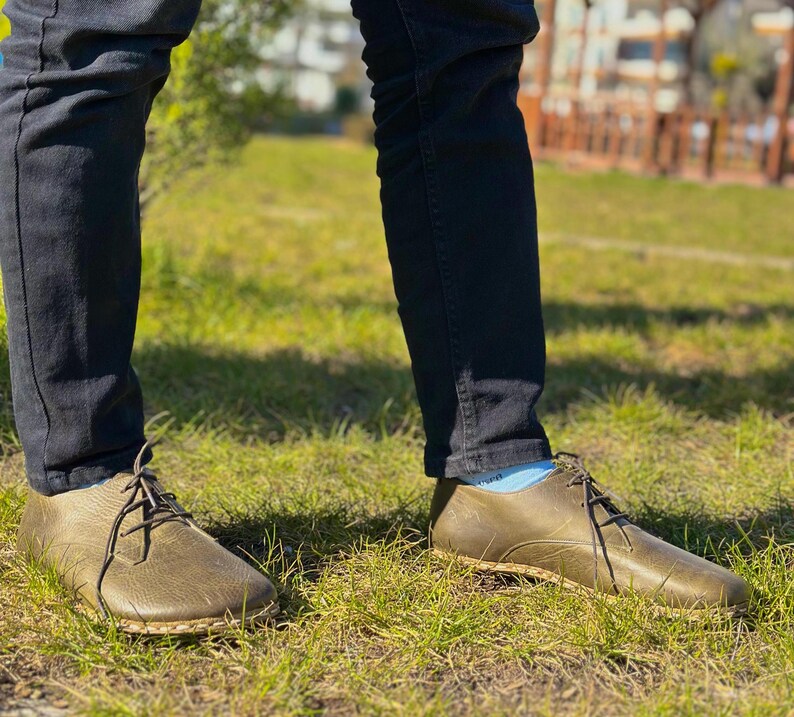 May include: A pair of olive green leather lace-up shoes with a rounded toe and a thick sole. The shoes are worn by a person wearing dark blue jeans.