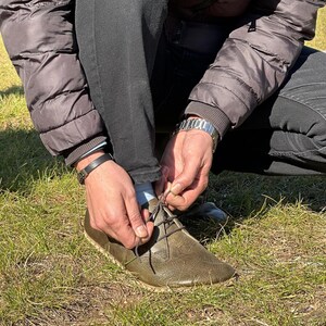 May include: A person is tying the laces of a brown leather shoe with a thick sole. The shoe has a lace-up design and is made of a textured material.