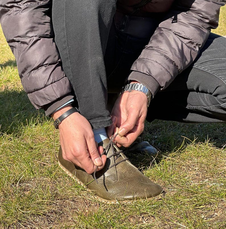 May include: A person is tying the laces of a brown leather shoe with a thick sole. The shoe has a lace-up design and is made of a textured material.
