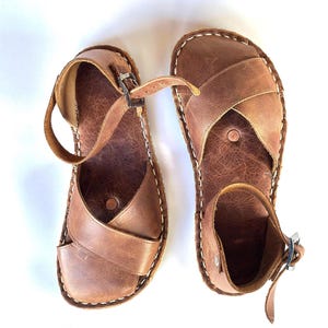Op de afbeelding: Een paar bruinleren sandalen met een kruisontwerp en enkelbandjes. De sandalen hebben witte stiksels rond de randen en zilveren gespen. De sandalen zijn ongeveer 25 cm lang.