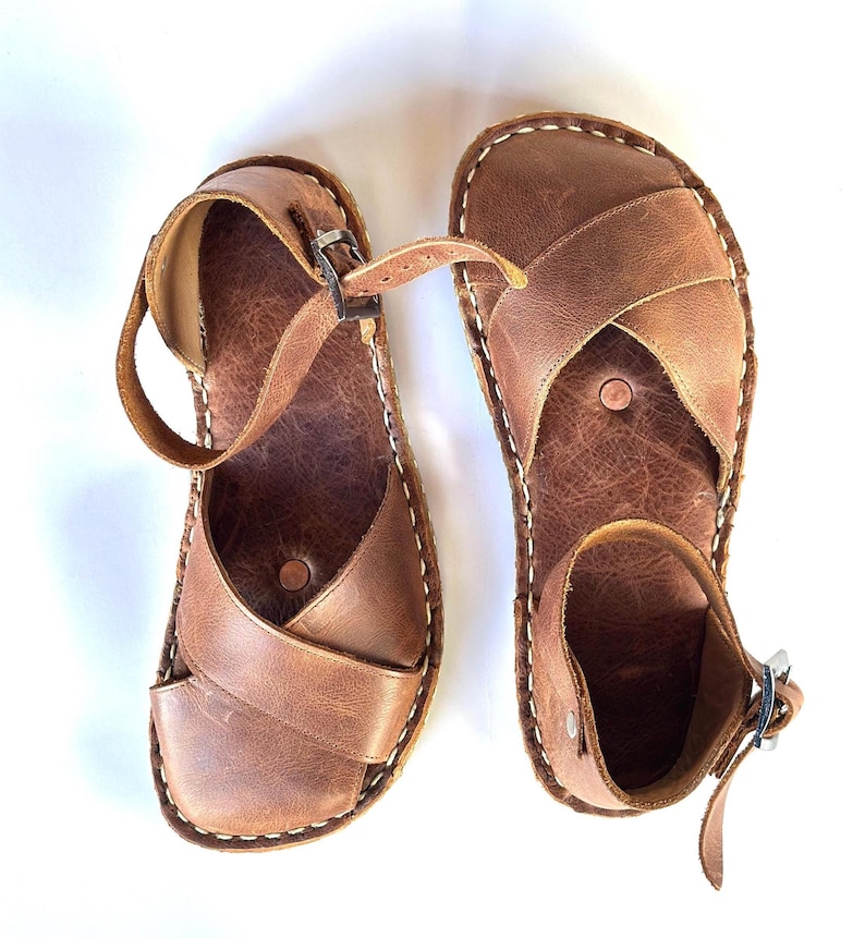 Op de afbeelding: Een paar bruinleren sandalen met een kruisontwerp en enkelbandjes. De sandalen hebben witte stiksels rond de randen en zilveren gespen. De sandalen zijn ongeveer 25 cm lang.