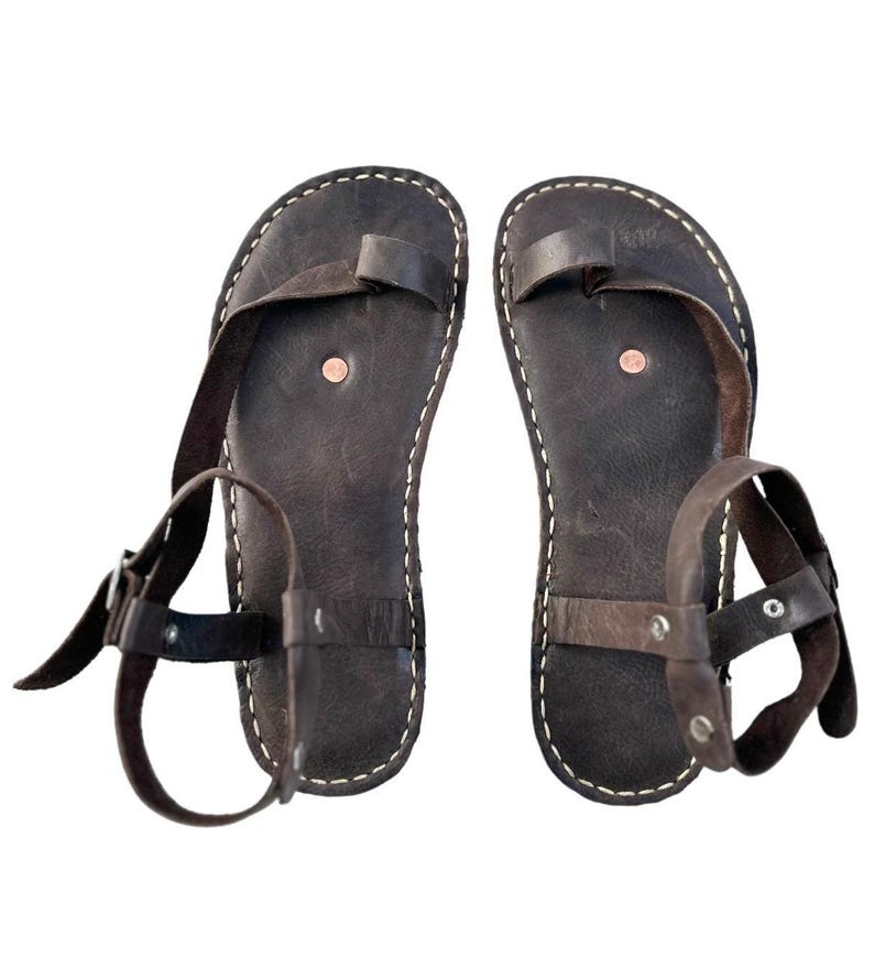 May include: A pair of brown leather sandals with ankle straps and buckles. The sandals have a simple design with a single strap across the toe and a strap around the ankle.