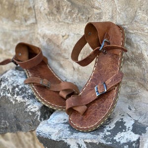 May include: A pair of brown leather sandals with buckles and straps. The sandals have a stitched edge and are made of a textured leather.