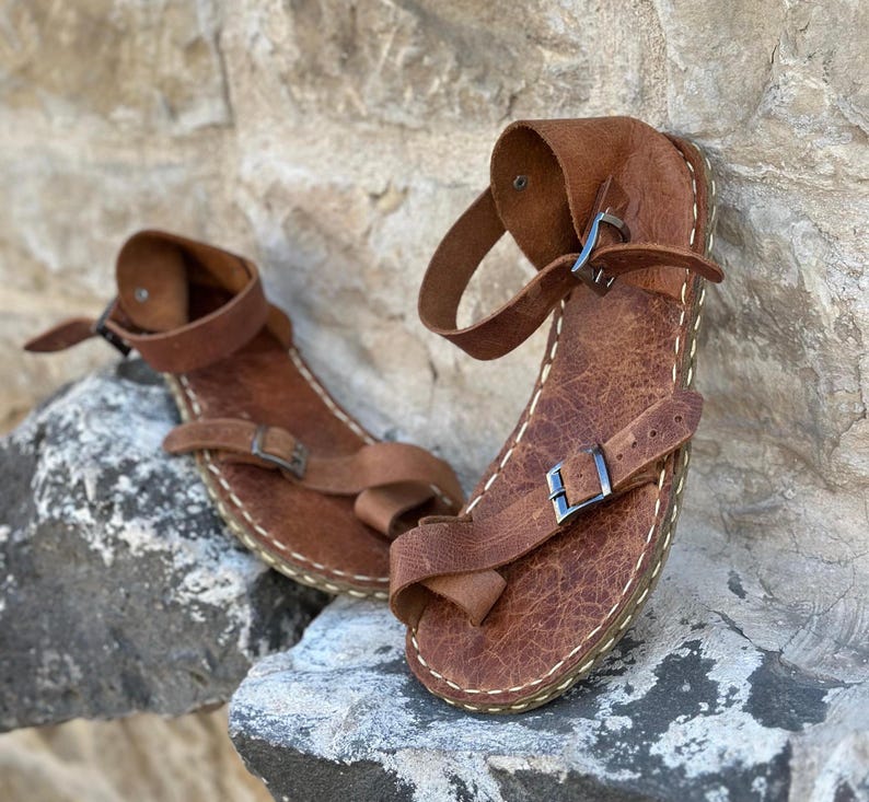 May include: A pair of brown leather sandals with buckles and straps. The sandals have a stitched edge and are made of a textured leather.