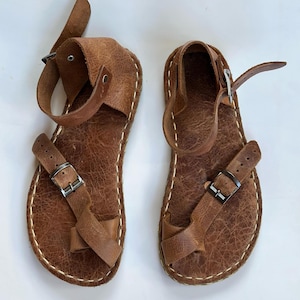 May include: A pair of brown leather sandals with buckles and straps. The sandals have a simple design and are made of high-quality leather.