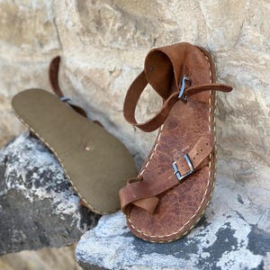 May include: Brown leather sandals with a buckle strap and a stitched edge. The sandals are on a rock surface.