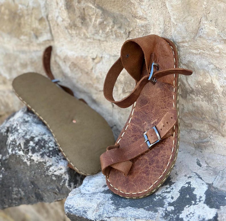 May include: Brown leather sandals with a buckle strap and a stitched edge. The sandals are on a rock surface.