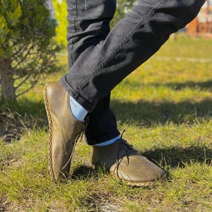 May include: A pair of brown leather shoes with laces. The shoes have a unique design with stitching around the edges. The shoes are worn by a person wearing jeans and white socks.