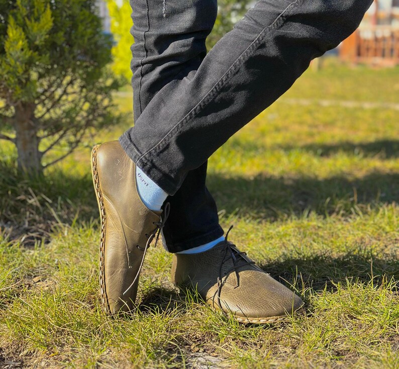 May include: A pair of brown leather shoes with laces. The shoes have a unique design with stitching around the edges. The shoes are worn by a person wearing jeans and white socks.