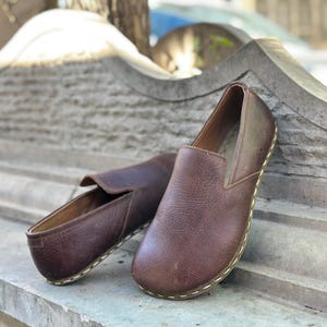 K&ouml;nnte beinhalten: Ein Paar braune Slipper aus Leder mit einem gen&auml;hten Rand. Die Schuhe befinden sich auf einer Steinoberfl&auml;che.