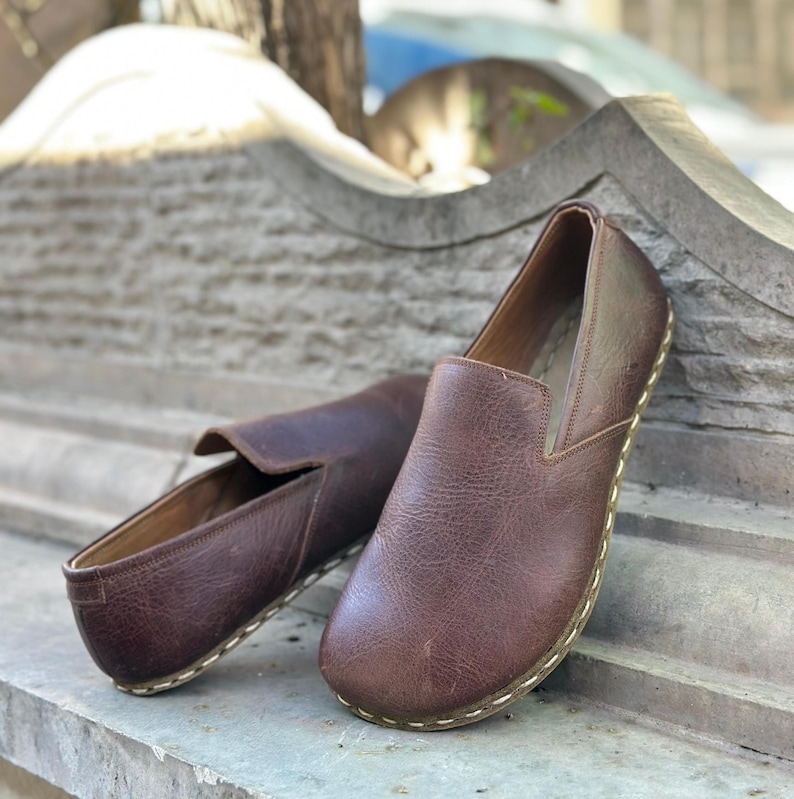 K&ouml;nnte beinhalten: Ein Paar braune Slipper aus Leder mit einem gen&auml;hten Rand. Die Schuhe befinden sich auf einer Steinoberfl&auml;che.