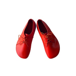 May include: A pair of bright red leather shoes featuring a rounded toe design, a low-cut silhouette, and thin matching red laces tied in a bow across the top. The shoes have a smooth, solid-colored finish and a simple, clean construction.
