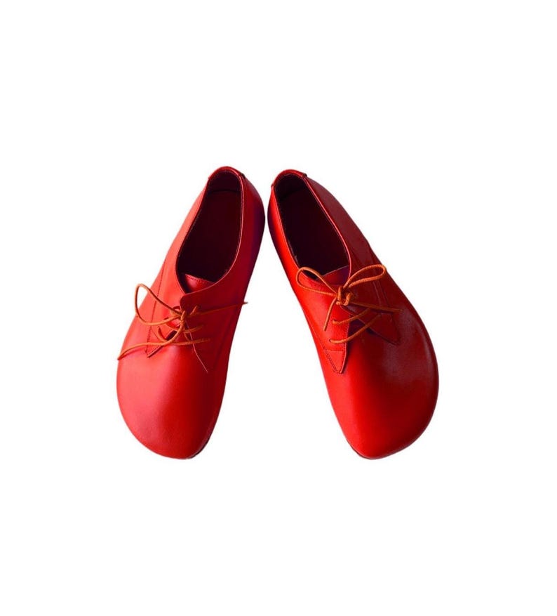 May include: A pair of bright red leather shoes featuring a rounded toe design, a low-cut silhouette, and thin matching red laces tied in a bow across the top. The shoes have a smooth, solid-colored finish and a simple, clean construction.