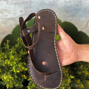 May include: Brown leather sandals with a single strap and a toe loop. The sandals have a white stitch detail around the edges.