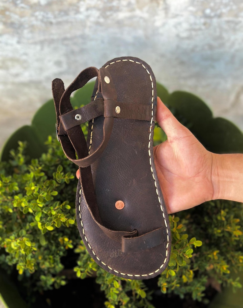 May include: Brown leather sandals with a single strap and a toe loop. The sandals have a white stitch detail around the edges.