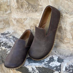 May include: Two pairs of brown leather slip-on shoes with a stitched edge. The shoes are on a rock surface.