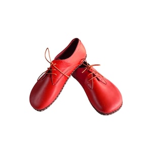 May include: A pair of bright red leather shoes featuring a rounded toe, a low-cut profile, and thin matching red laces. The shoes have visible stitching along the sole edge and a smooth, matte finish, presented against a plain white background.