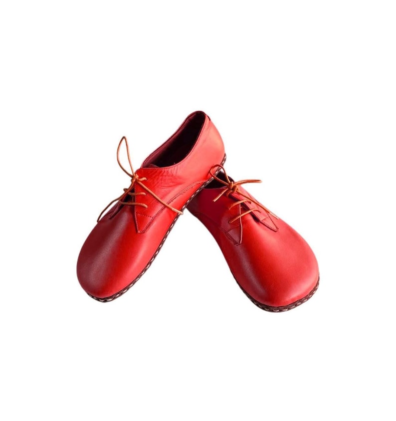 May include: A pair of bright red leather shoes featuring a rounded toe, a low-cut profile, and thin matching red laces. The shoes have visible stitching along the sole edge and a smooth, matte finish, presented against a plain white background.