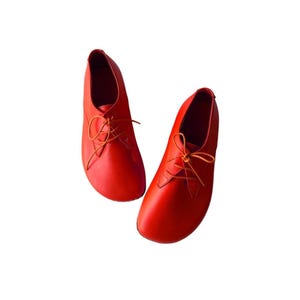 May include: A pair of vibrant orange leather shoes featuring a rounded toe design, a low-cut ankle profile, and thin matching orange laces threaded through two eyelets on each side. The shoes have a smooth, matte finish and a minimalist construction.