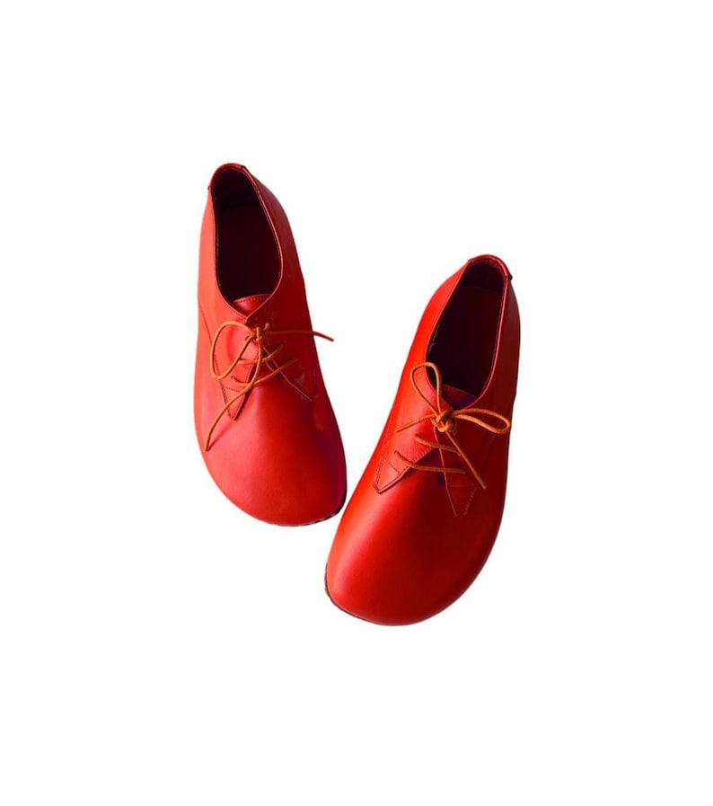 May include: A pair of vibrant orange leather shoes featuring a rounded toe design, a low-cut ankle profile, and thin matching orange laces threaded through two eyelets on each side. The shoes have a smooth, matte finish and a minimalist construction.