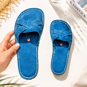 Puede incluir: Un par de sandalias de cuero azul con un diseño de tiras cruzadas en la parte superior, costuras blancas en contraste a lo largo de los bordes y un pequeño detalle de remache circular de color cobre centrado en la plantilla de cada sandalia.