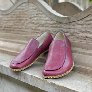 Puede incluir: Un par de zapatos de cuero burdeos de vestir sin cordones con un acabado texturizado sutil. Los zapatos tienen una costura cosida alrededor del borde de la suela y una suela de color marr&oacute;n claro.