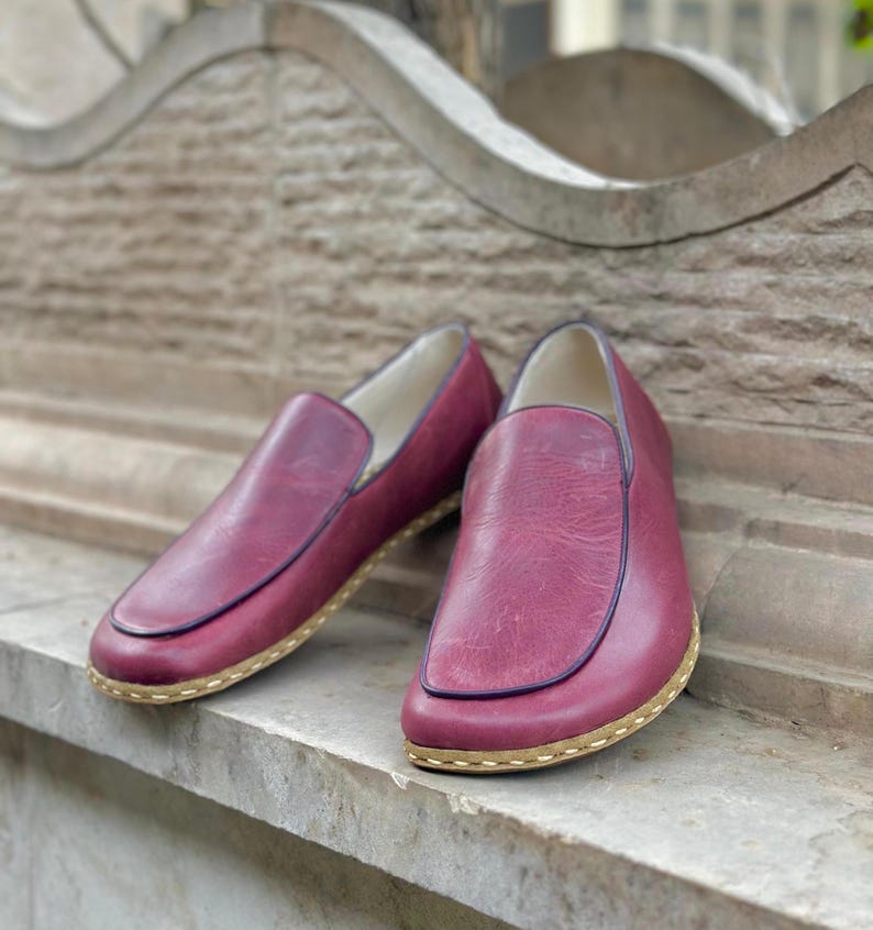 Puede incluir: Un par de zapatos de cuero burdeos de vestir sin cordones con un acabado texturizado sutil. Los zapatos tienen una costura cosida alrededor del borde de la suela y una suela de color marr&oacute;n claro.