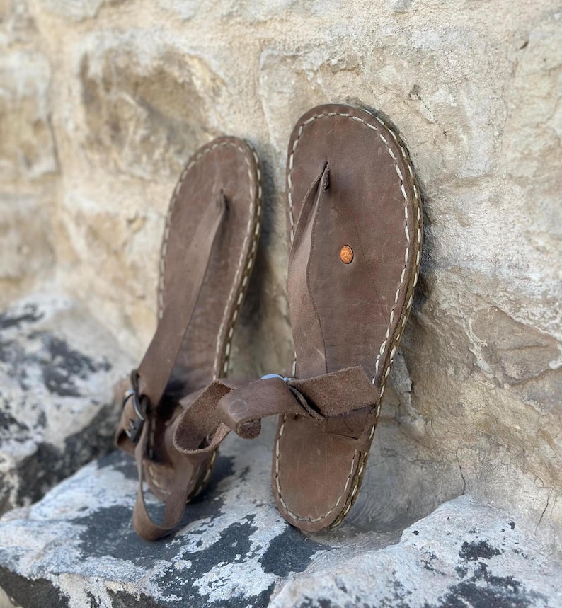 Puede incluir: Un par de sandalias de cuero marr&oacute;n con una correa de estilo thong y un detalle de costura blanca alrededor de los bordes.