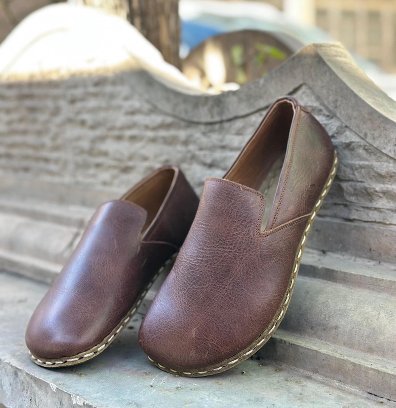 K&ouml;nnte beinhalten: Ein Paar braune Slipper aus Leder mit einem gen&auml;hten Rand. Die Schuhe befinden sich auf einer Steinoberfl&auml;che.