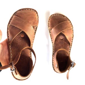 Op de afbeelding: Een paar bruin lederen sandalen met een kruisontwerp aan de bovenkant. De sandalen hebben een enkelbandje en zijn gestikt met wit garen. De sandalen worden op een witte achtergrond getoond.