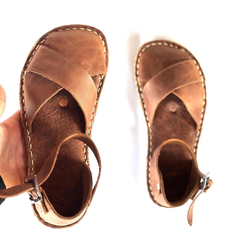 Op de afbeelding: Een paar bruin lederen sandalen met een kruisontwerp aan de bovenkant. De sandalen hebben een enkelbandje en zijn gestikt met wit garen. De sandalen worden op een witte achtergrond getoond.