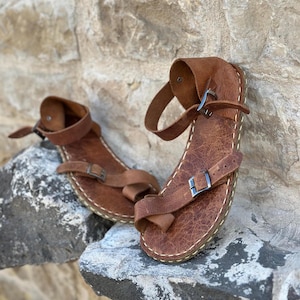 May include: A pair of brown leather sandals with buckles and straps. The sandals have a stitched edge and are sitting on a rocky surface.